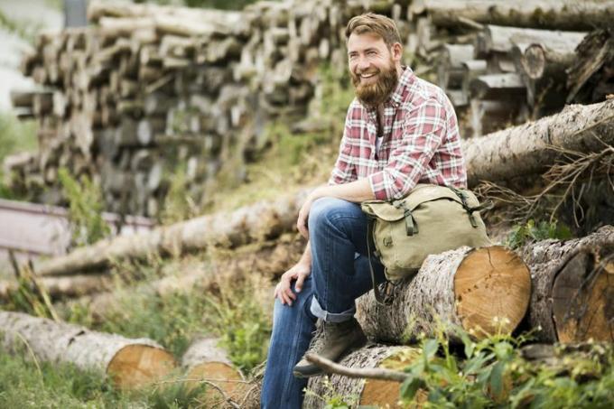 Hombre barbudo riendo sentado sobre una pila de madera recién cortada