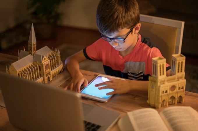 صبي صغير يرتدي نظارات يجلس على الطاولة مع جهاز لوحي محاط بنماذج المباني