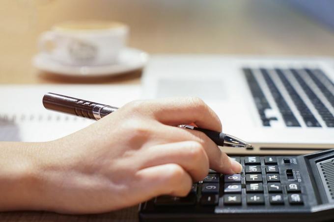 Ženska ruka radi s kalkulatorom, poslovnim dokumentom i prijenosnim računalom.