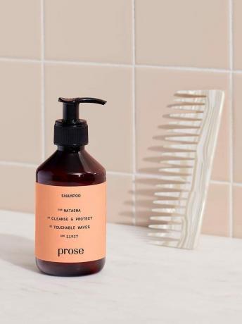 El producto de champú se encuentra en el mostrador de un baño junto a un peine apoyado contra una pared de azulejos rosas.