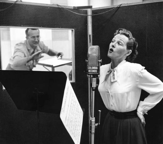 CIRCA 1955: Die amerikanische Pop- und Jazzsängerin Jo Stafford (1917-2008) singt in einem Aufnahmestudio in ein Mikrofon, während ihr Ehemann, der Arrangeur Paul Weston, aus dem Nebenraum dirigiert.