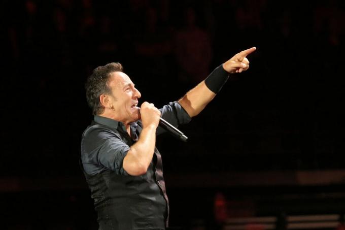 Bruce Springsteen profilde karanlık bir sahnede performans sergiliyor.