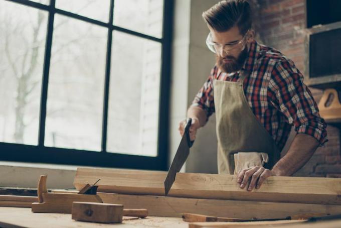 jeune ébéniste élégant avec lunettes et coiffure. Fort bel artisan tenant une scie et du bois vierge sur le lieu de travail