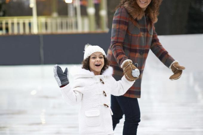 Mergina čiuožia ant ledo su mama, susikibusi už rankų žiūri į kamerą šypsodamasi