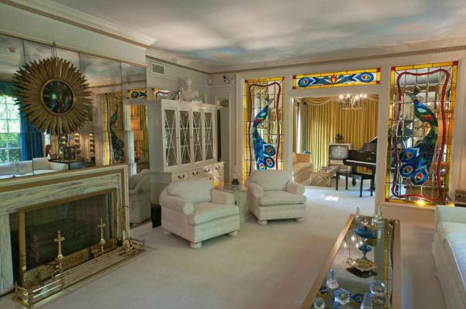 Şöminenin üzerinde aynalar, beyaz duvarlı mobilyalar, kuyruklu piyanolu müzik odasına açılan vitraylı açık kapı ile Graceland Mansion oturma odasının başka bir görünümü
