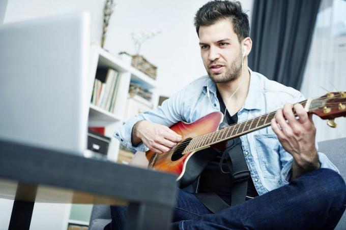 Млад мъж у дома, седнал на дивана, свирещ на китара и гледащ лаптопа