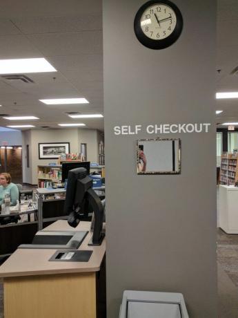 Bir kütüphanede çekilmiş bir fotoğraf. Duvarda bir ayna var. Aynanın üzerinde " Kendi Kendine Ödeme" yazan bir işaret var.